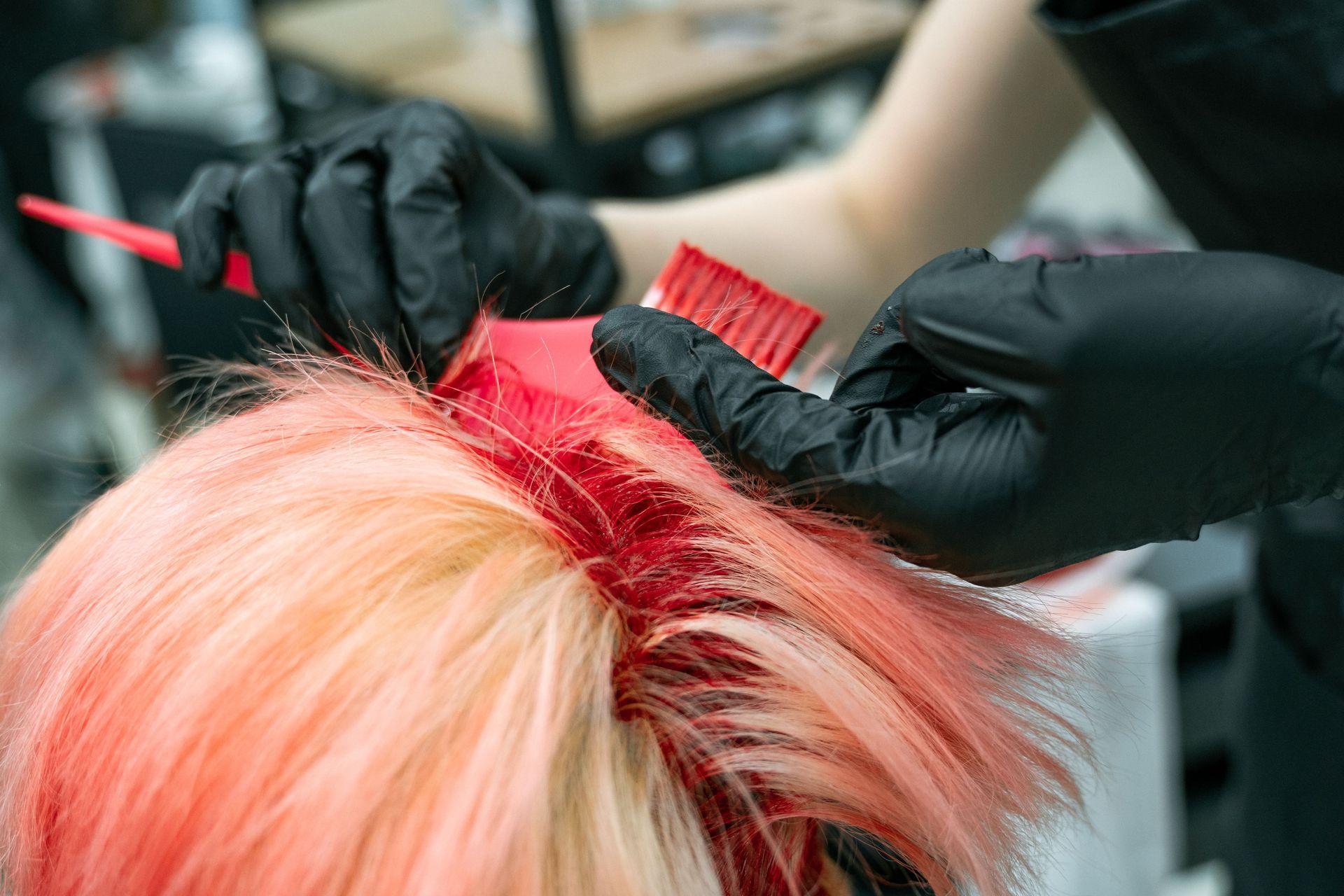 Gloved hands part bright red hair while coloring the roots in a salon