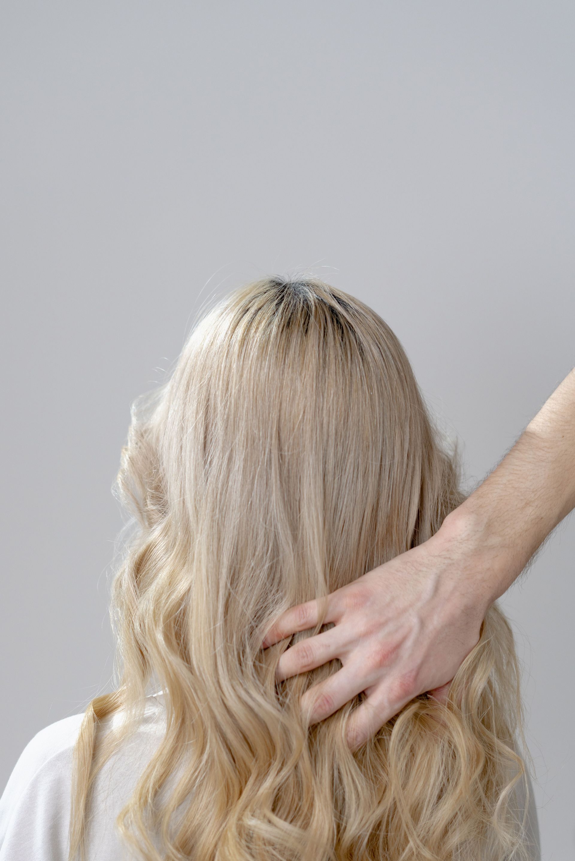 Hand touching the back of a blonde person’s wavy hair against a plain gray background