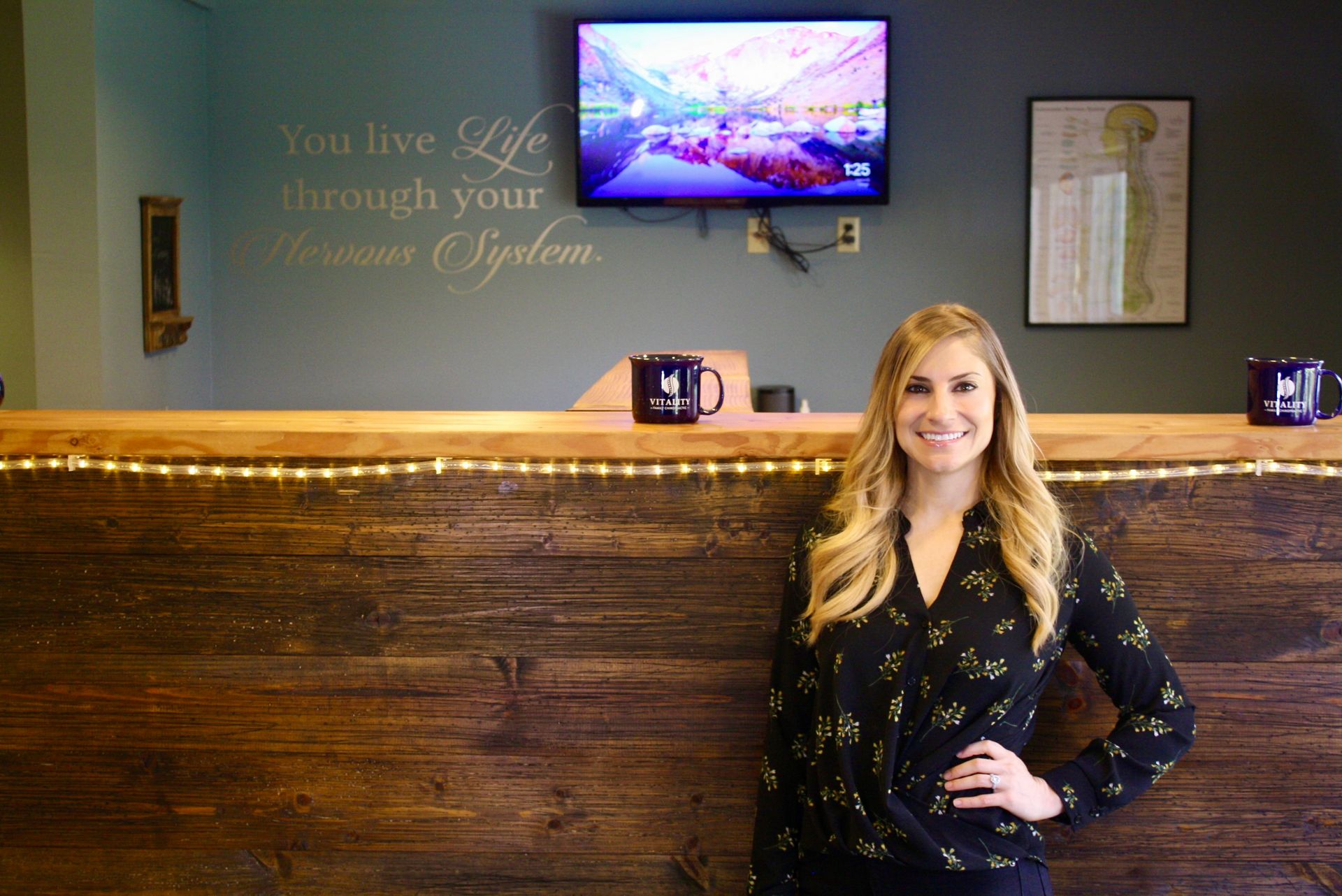 Woman standing behind a wood reception desk in a lobby with wall art and a TV screen above.