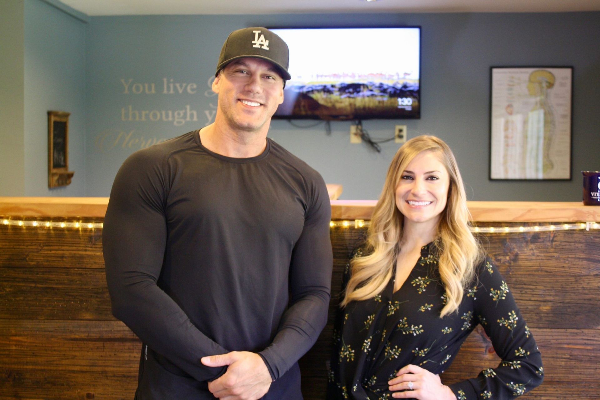 Two smiling people standing in a warmly lit office lobby, with framed wall art and a TV behind them