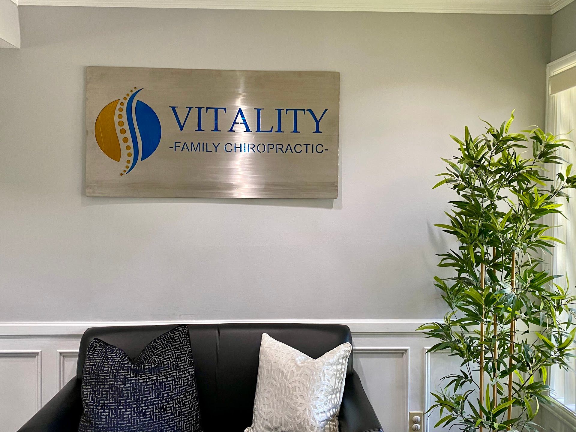 Vitality Chiropractic sign above a black sofa with decorative pillows and a tall bamboo plant