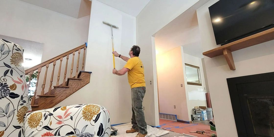 Person using a paint roller to paint a white wall in a living room near stairs and a fireplace.