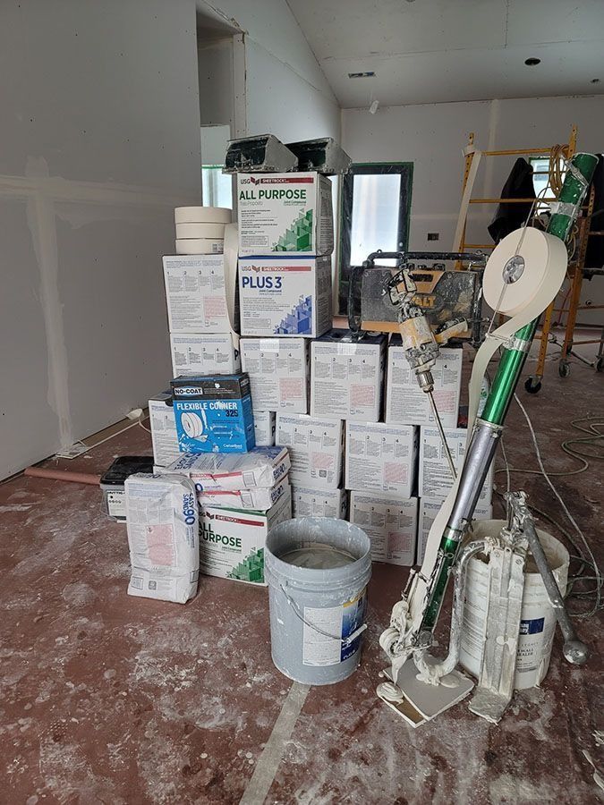 Stacked paint and supply boxes in a room under renovation, with tools and a bucket on a dusty floor.