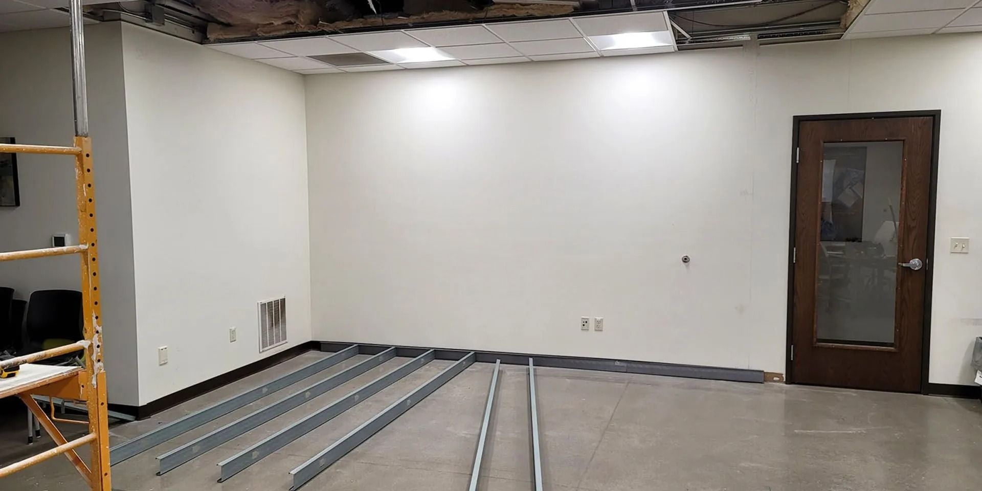 Empty room under renovation with exposed floor beams, white walls, and a brown door on the right