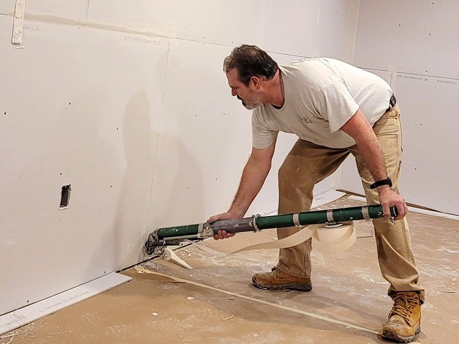 Man applying green paint or sealant along a wall base in a room with plastic-covered floor
