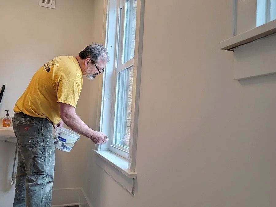 Person painting a window frame inside a room, wearing a yellow shirt and jeans.