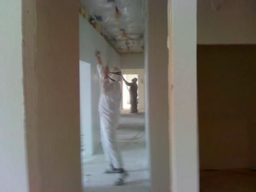 Person painting a hallway interior, wearing white coveralls and reaching toward the ceiling.