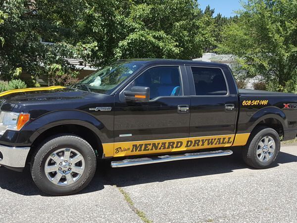 Black and gold Menard Drywall pickup truck parked on a driveway with trees in the background