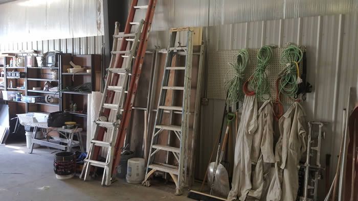Ladders, tools, and hanging work clothes stored along a corrugated metal wall in a workshop.