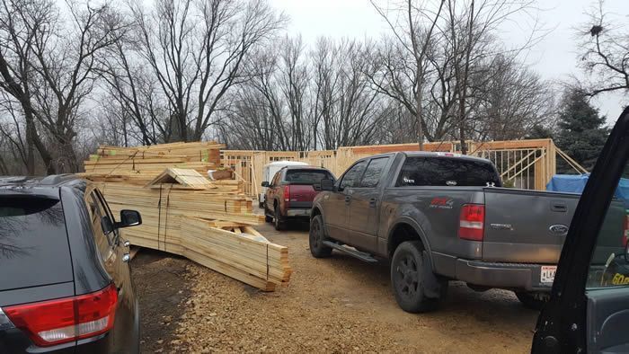 Pickup trucks parked beside stacks of lumber at a construction site on a muddy, wooded lot