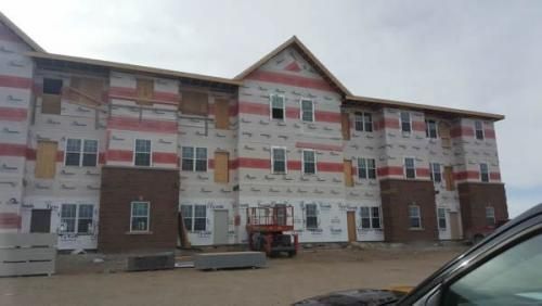 Three-story apartment building under construction, wrapped in white and red sheathing with a lift in front.