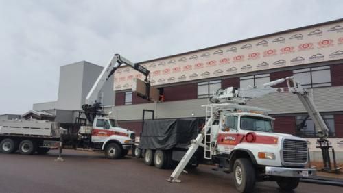 Two crane trucks lifting a large wall panel at a construction site near a warehouse.