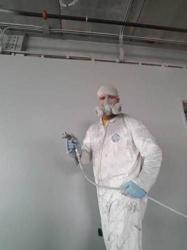 Person in white protective suit and mask spray-painting a wall in an industrial interior.