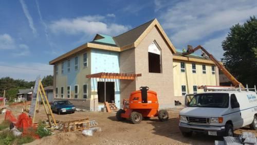 House under construction with workers, scaffolding, and trucks in a dirt lot