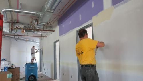 Two workers painting a hallway wall, one on a lift and one on the floor, in a building under renovation