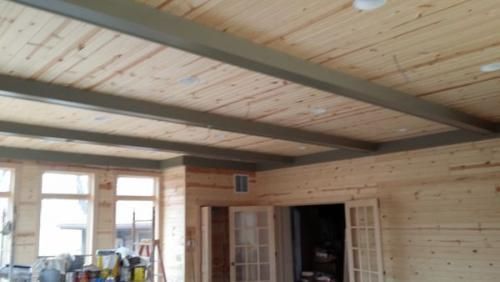 Wood-paneled room with exposed dark ceiling beams, windows, and an open doorway; tools and supplies are visible.