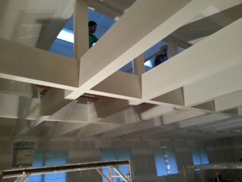 Ceiling view of white steel beams in an industrial interior with a few workers visible above.