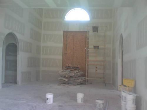 Unfinished room with drywall, arched window, central wooden door, and buckets on the floor.