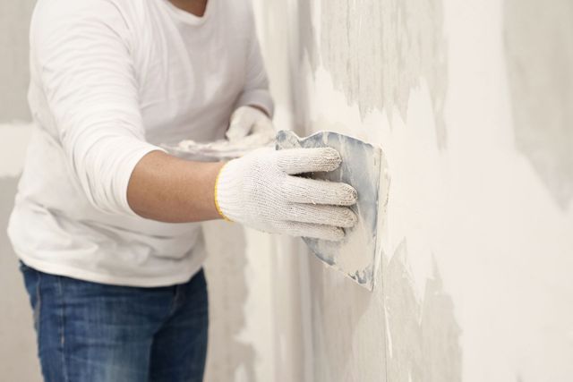 Person in white shirt and gloves smoothing wet plaster on a wall with a trowel