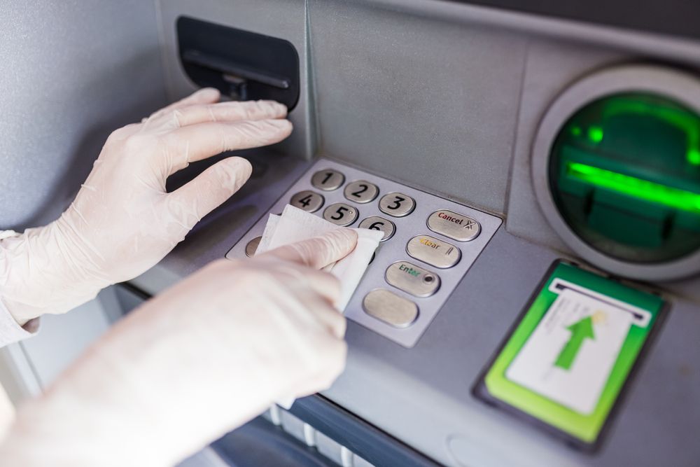 Gloved hands using an ATM keypad with a card inserted and green-lit slot, likely entering a PIN