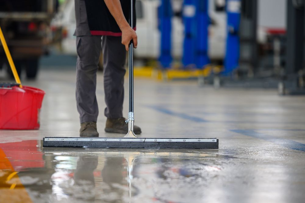Worker mopping a wet factory floor with a squeegee; red bucket nearby.