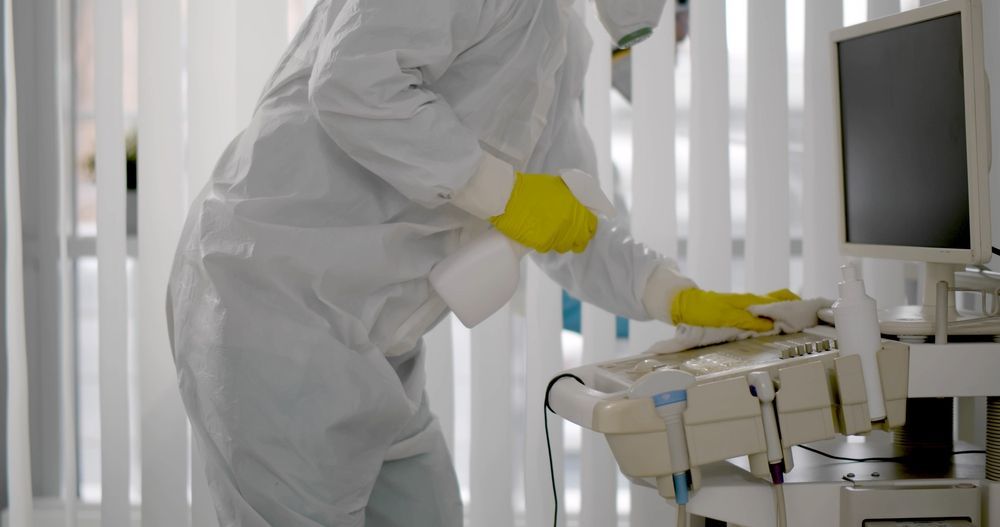 Person in white protective suit and yellow gloves cleaning lab equipment beside a monitor