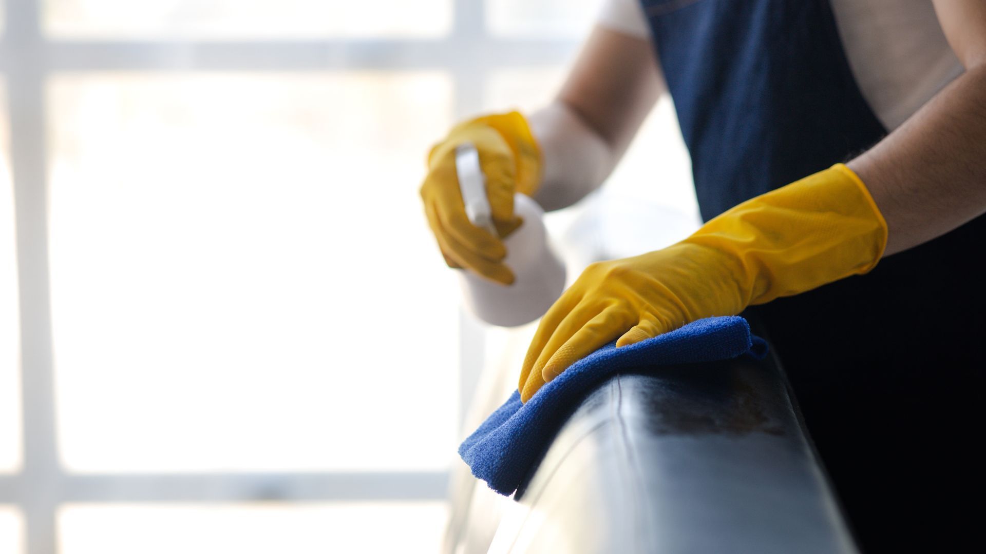 Person wearing yellow gloves wiping a chair with a blue cloth in a bright room