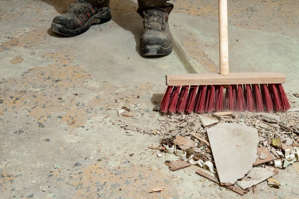 A broom sweeps broken concrete debris from a dusty floor near work boots.