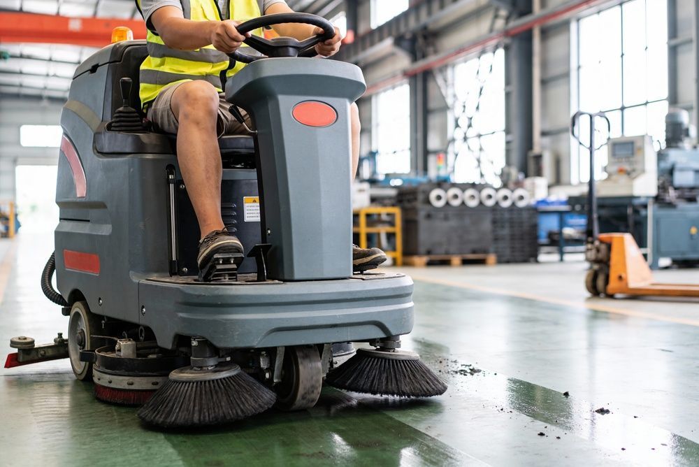 Industrial floor scrubber cleaning a warehouse floor, with a worker operating it.