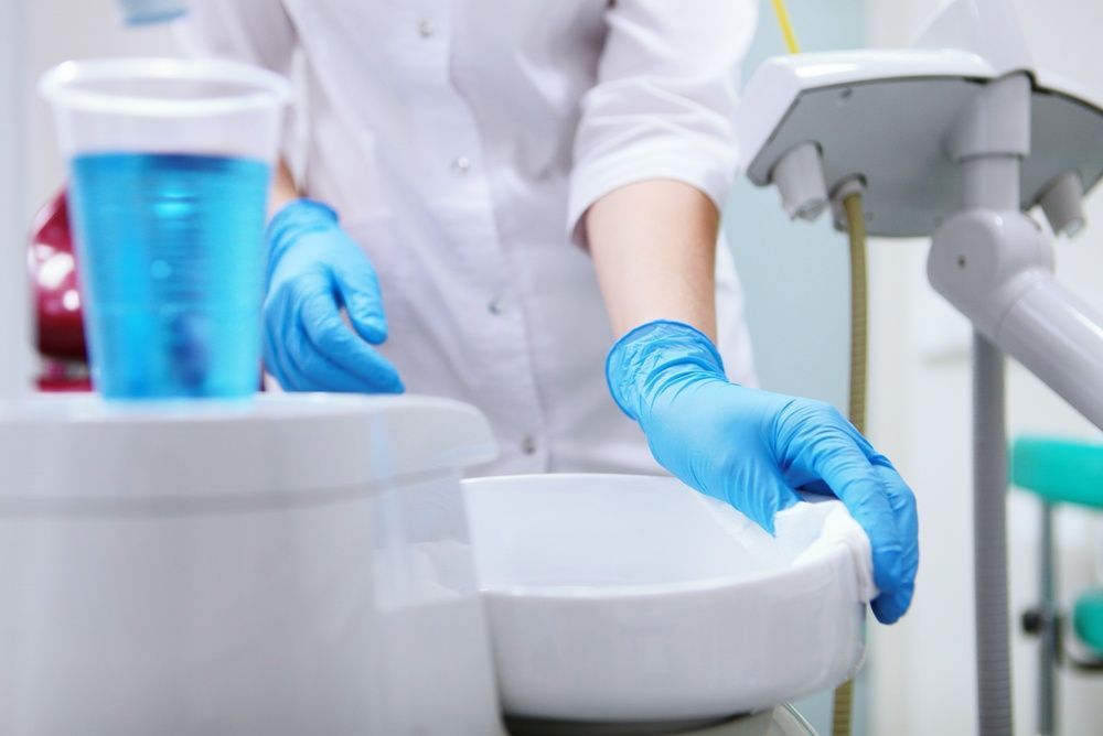 Gloved worker cleaning a white dental chair with a blue cup and equipment nearby