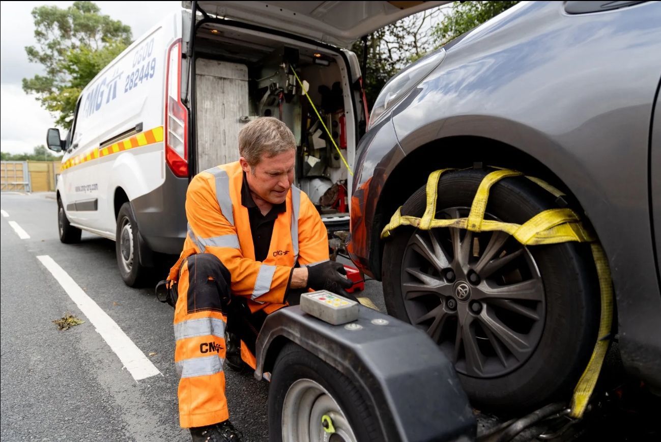 Tow truck operator securing a gray car to a trailer. Operator in orange workwear, van in background.