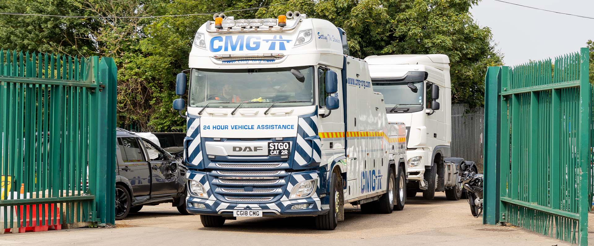 White cargo truck in a green gate entrance, with parked vehicles and a concrete yard behind it