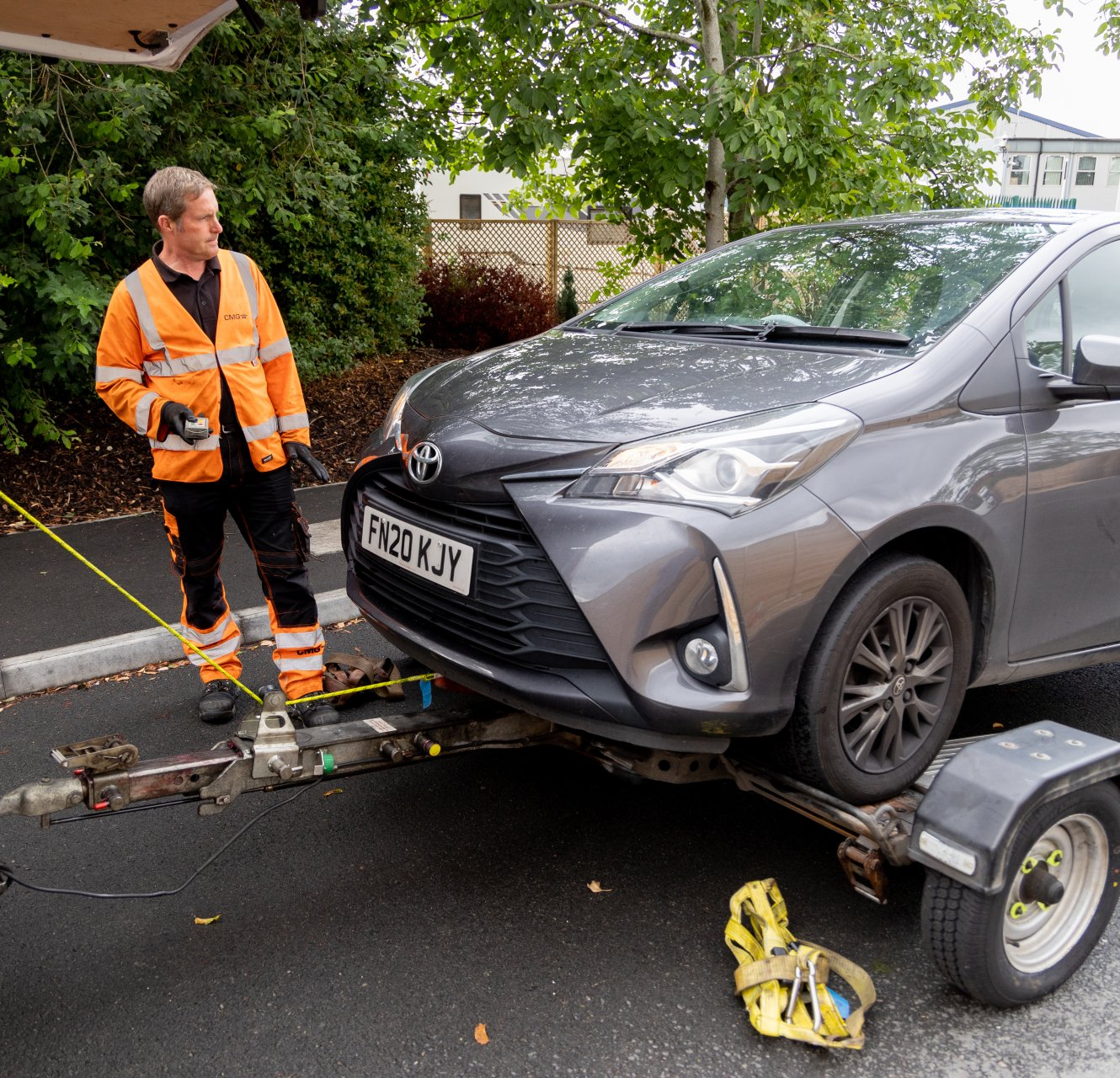 Man in safety vest loading a gray Toyota Yaris onto a trailer.