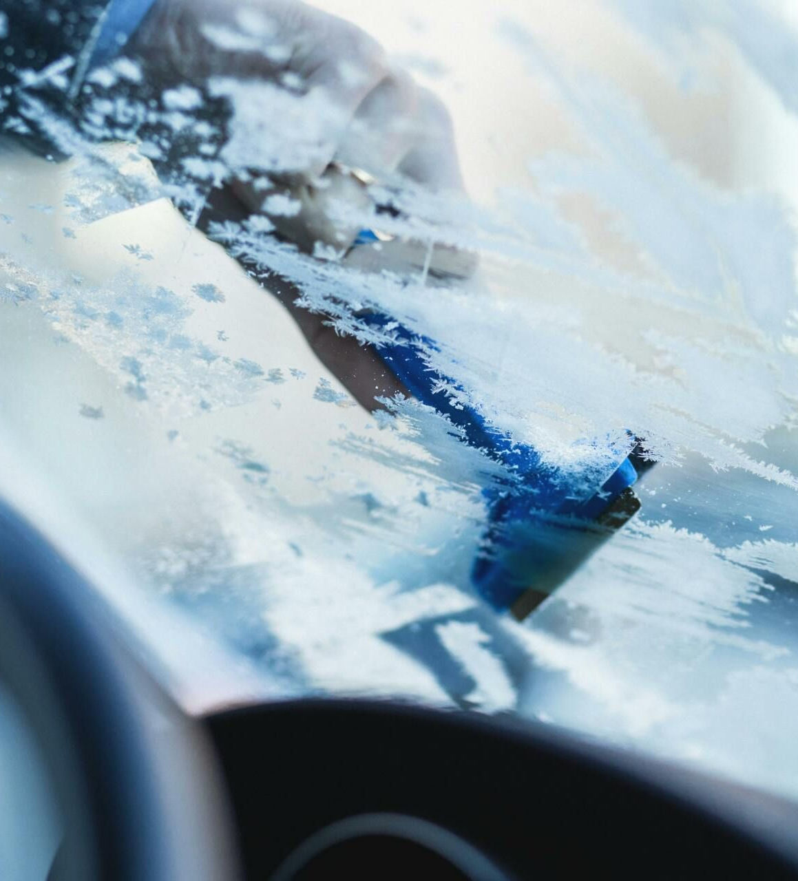 Person scraping ice from a car windshield with a blue scraper on a cold, snowy day.