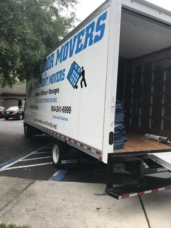 Moving truck with open back door, parked on a sidewalk. White truck with blue lettering for Door Mover company.