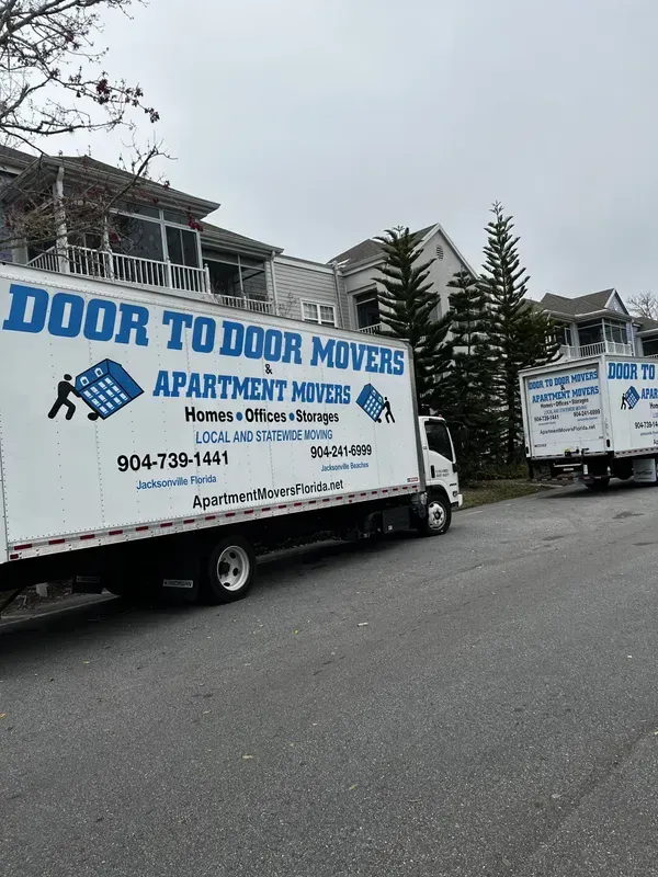 Two movers in blue overalls push a large wrapped object on a hand truck in a kitchen, boxes nearby. Two movers in blue overalls push a large wrapped object on a hand truck in a kitchen, boxes nearby.