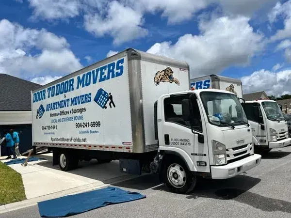 Moving trucks parked near a building; one has a logo reading Door to Door Movers. Two people are working.