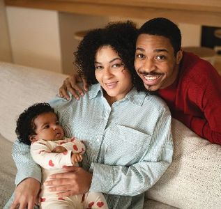 Family of three, smiling. Woman holding baby, man with arm around her. Inside, on a couch.