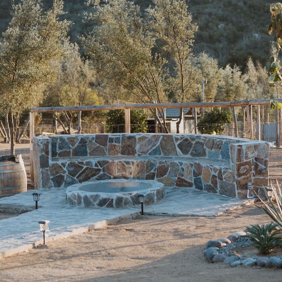 Fogón exterior de piedra y bancos curvos en un jardín rústico, con árboles al fondo