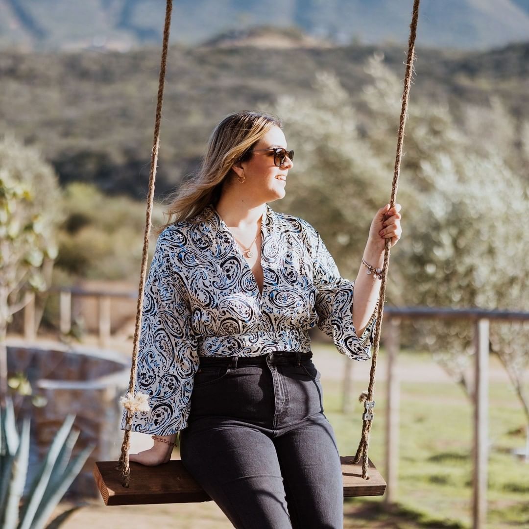 Mujer con gafas de sol sentada en un columpio al aire libre, con una blusa estampada y pantalones negros