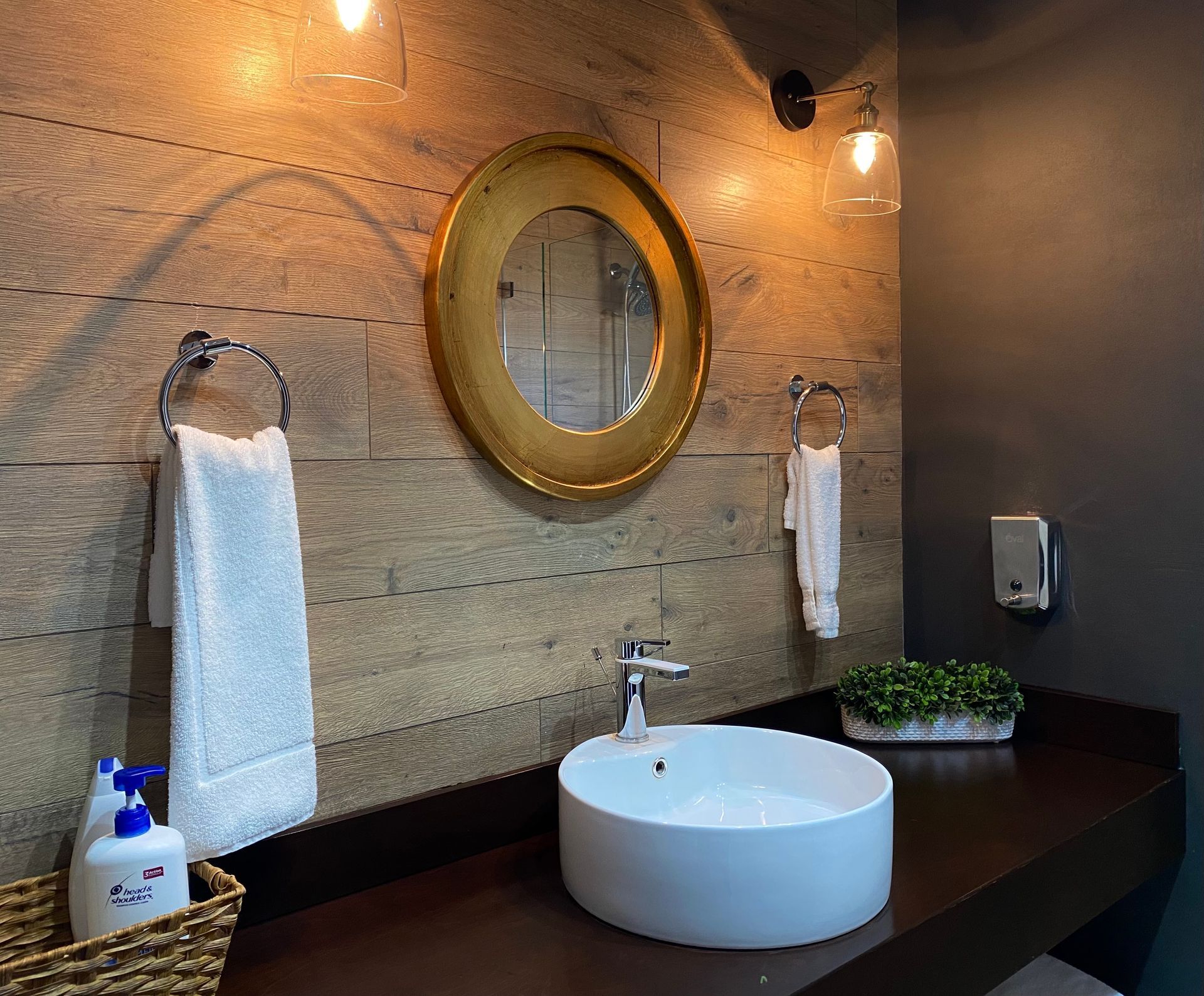 Mueble de baño moderno con espejo redondo, lavabo blanco, encimera oscura y toallas colgadas en la pared