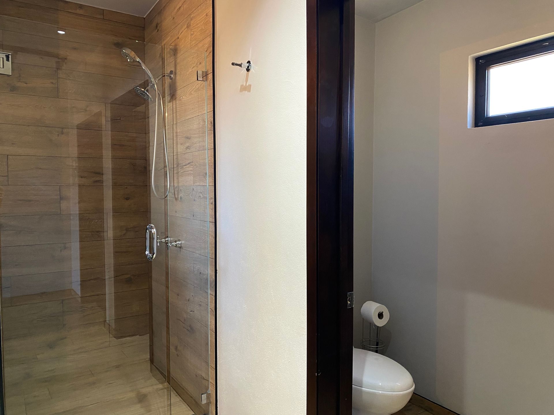 Baño con ducha de cristal, azulejos beige e inodoro visible a través de la puerta abierta.