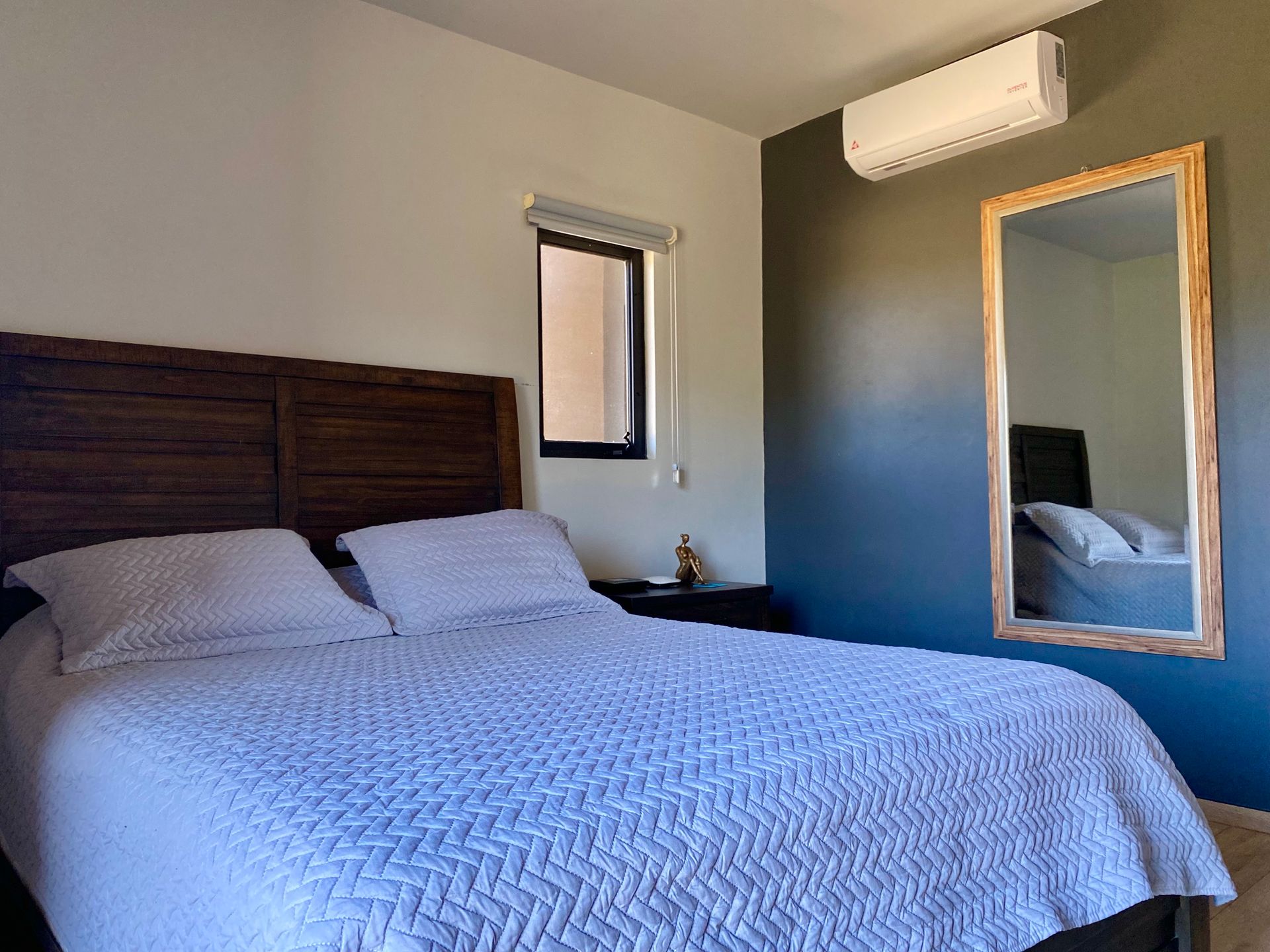 Dormitorio moderno con una cama de madera oscura, ropa de cama blanca, un espejo de pared y aire acondicionado.