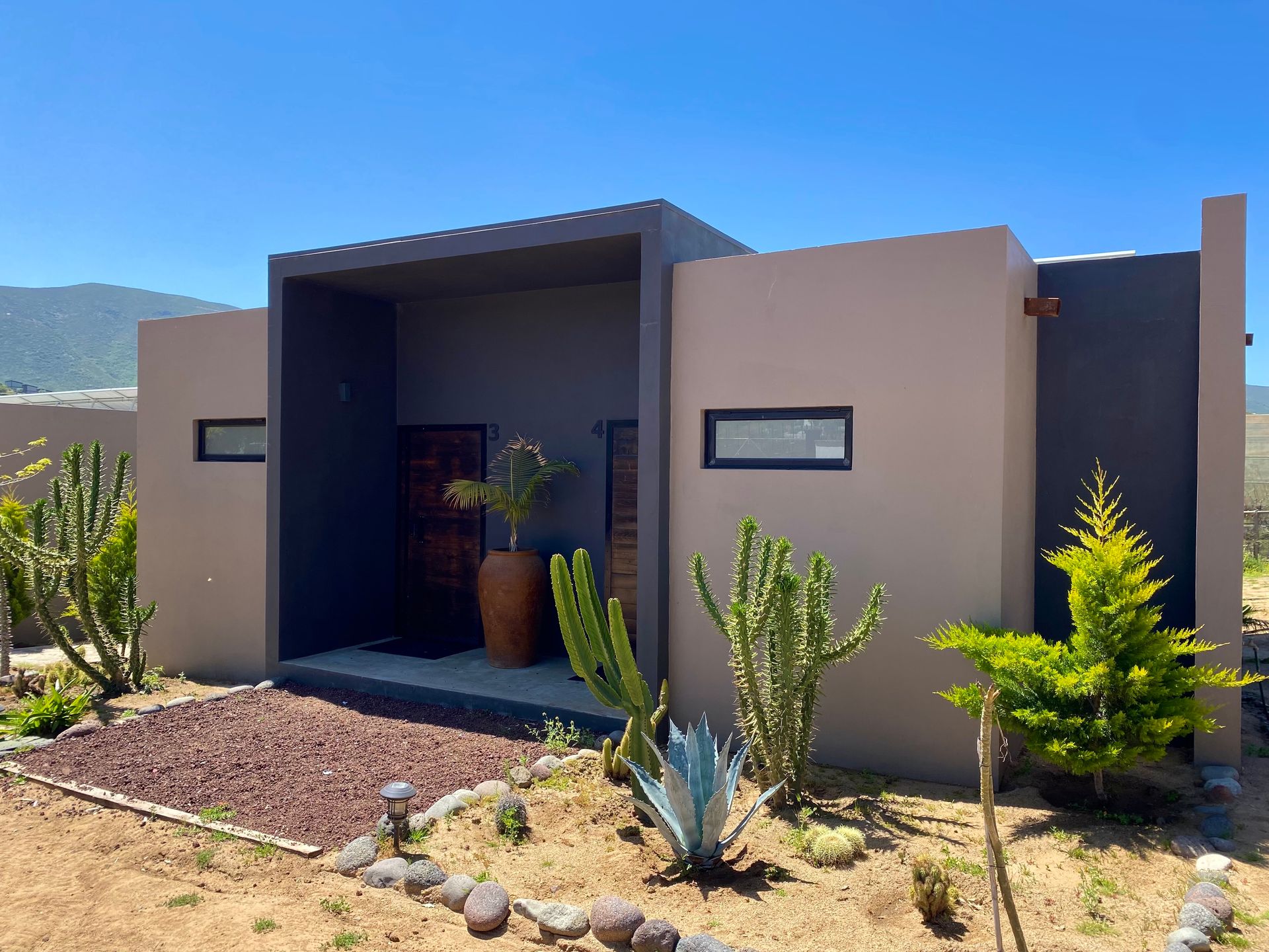 Casa beige moderna con una entrada oscura y un jardín desértico bajo un cielo despejado
