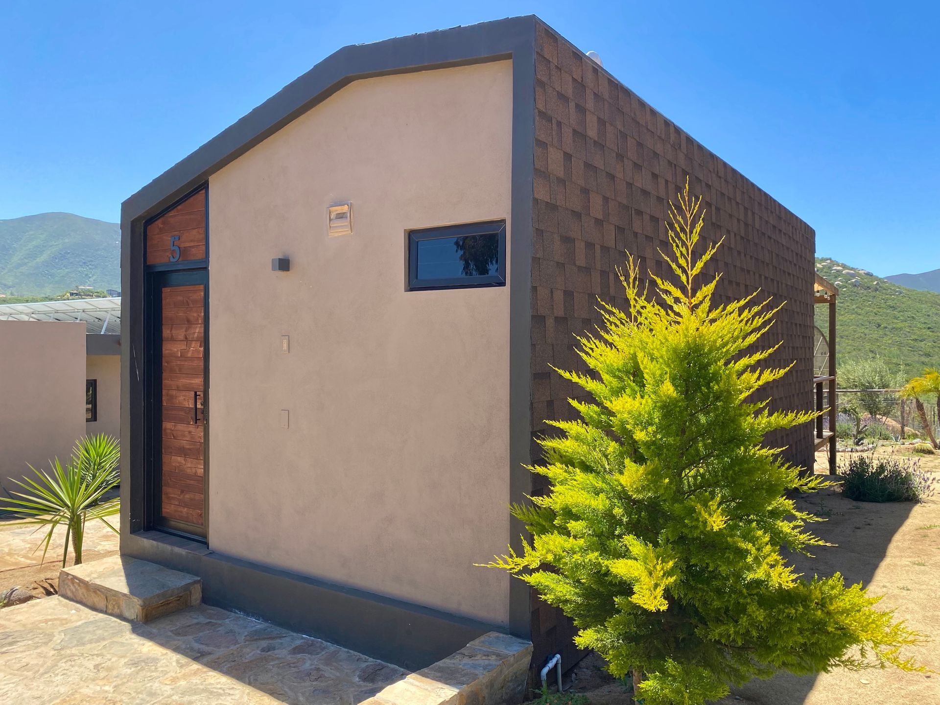 Pequeño edificio de estuco beige con molduras oscuras, una ventana estrecha y un arbusto de un verde intenso delante.