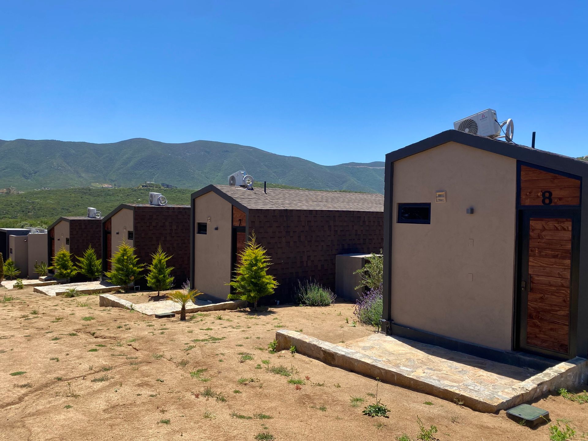 Fila de pequeñas cabañas beige con tejados marrones en un patio árido, con montañas verdes al fondo bajo un cielo despejado