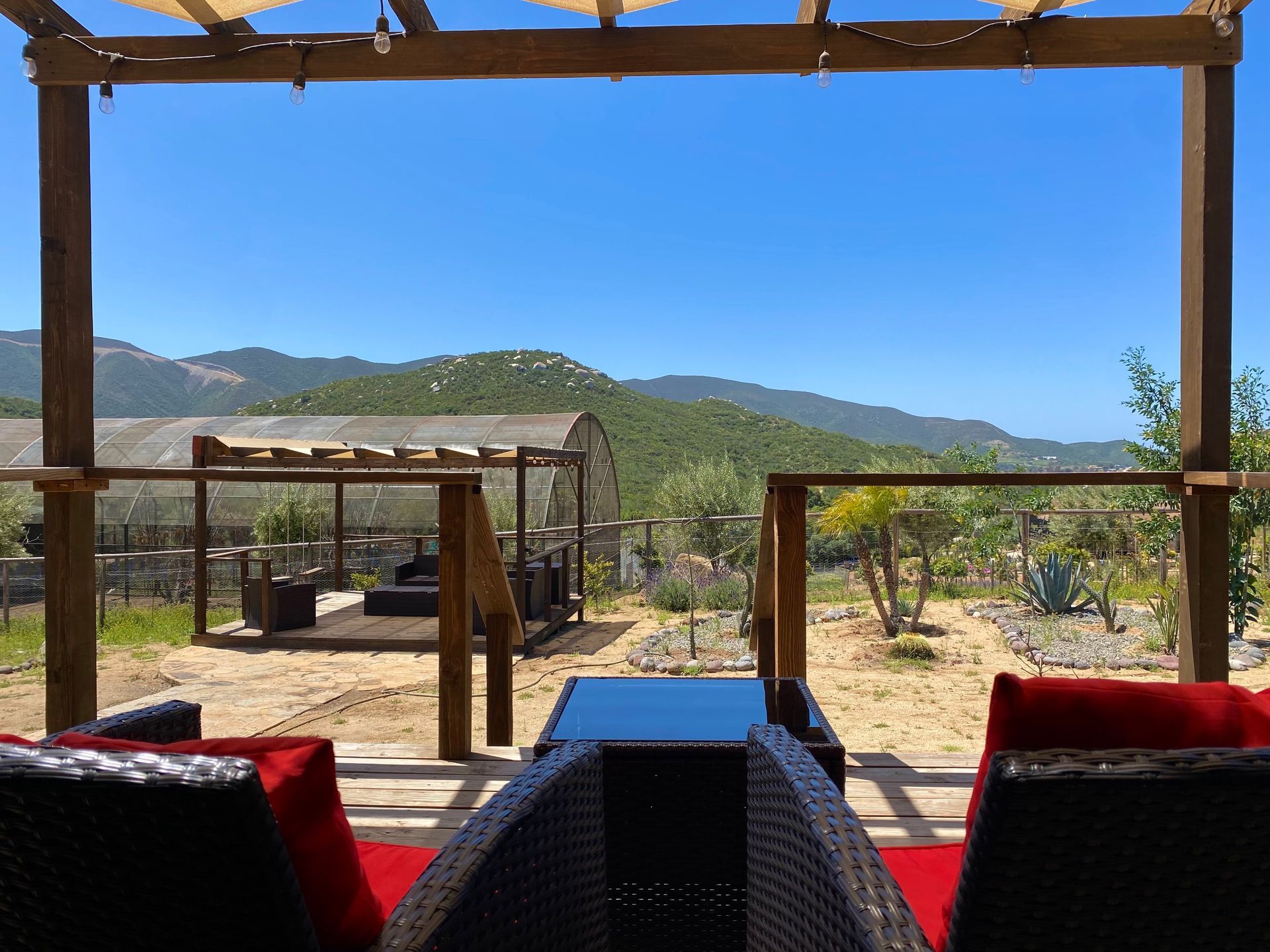 Patio al aire libre con pérgolas de madera, cojines rojos y vistas a las montañas bajo un cielo azul despejado