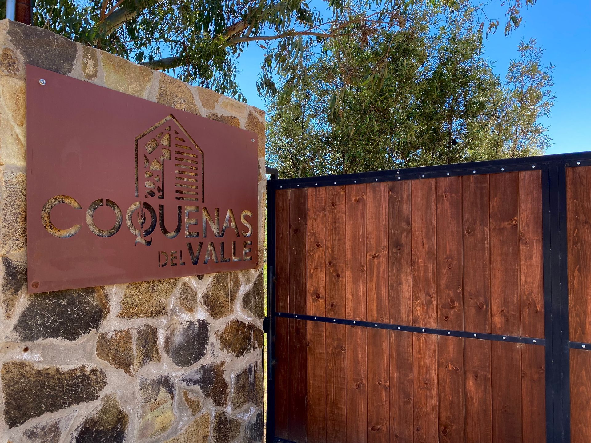Cartel de “Coquenas del Valle” junto a una puerta de madera y un muro de piedra bajo un cielo azul