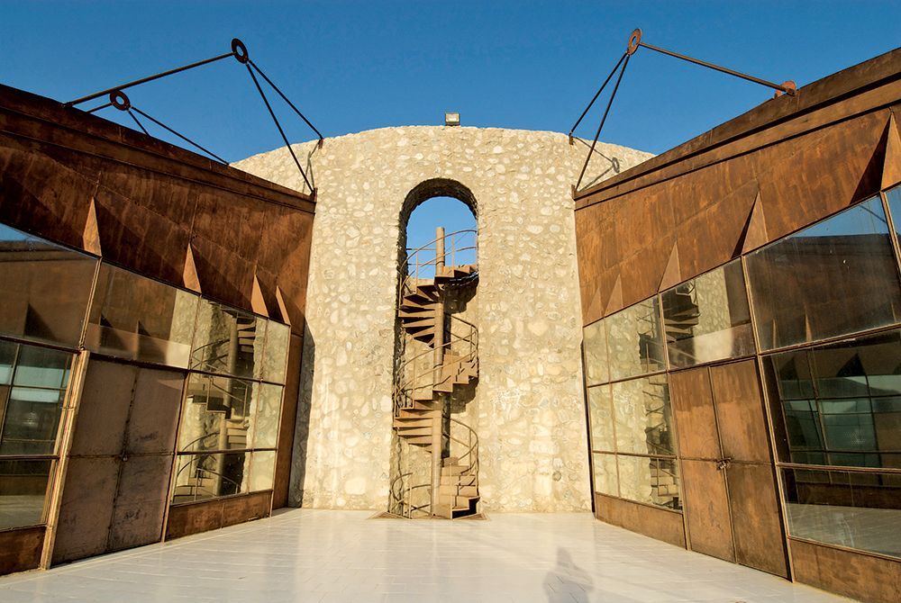 Entrada moderna de edificio con paredes de cristal espejado y un arco de piedra que enmarca una puerta estrecha