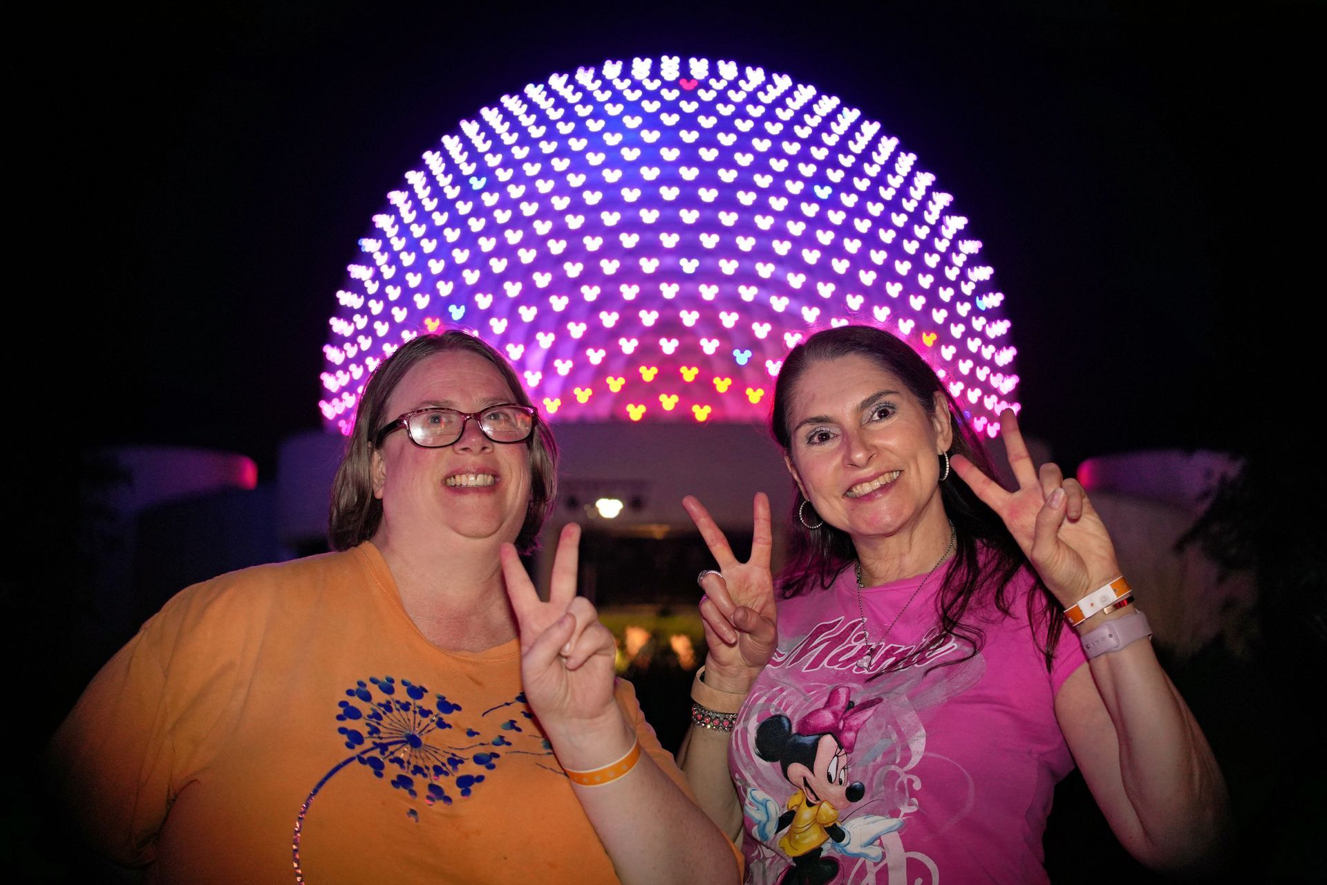 Two people smiling and flashing peace signs in front of a glowing purple geodesic dome at night